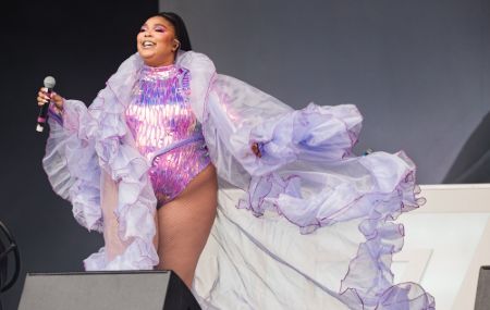 lizzo on the stage holding a mic on a holographic purple body suit with a sheer purple robe over her. lizzo has a purple makeup and high ponytail. 